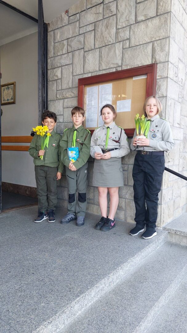 Four children in scout uniforms stand on stone steps outside a building, holding flowers and a card.