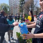 Young woman hands out a blue gift carton and a potted plant with yellow flowers at an outdoor community event.