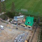 Aerial view of a damaged building with green and turquoise roofs beside a soccer field; firefighters on site and construction equipment nearby.