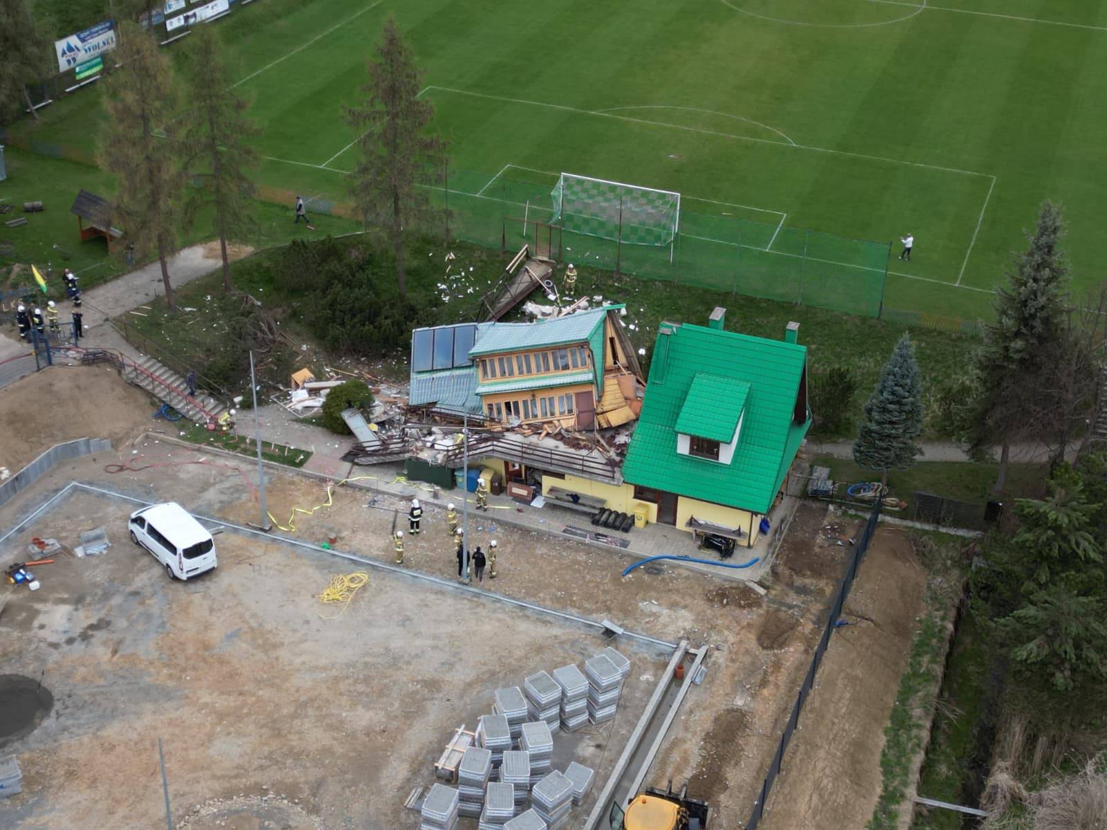 Aerial view of a damaged building with green and turquoise roofs beside a soccer field; firefighters on site and construction equipment nearby.