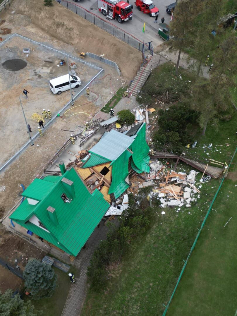 A green-roofed house heavily damaged with a collapsed roof and debris scattered in the yard; firefighters and a white van present at the site.