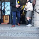 Man in a dark coat hands a bright yellow flower to a student at a doorway during a ceremony, with others nearby on the steps.