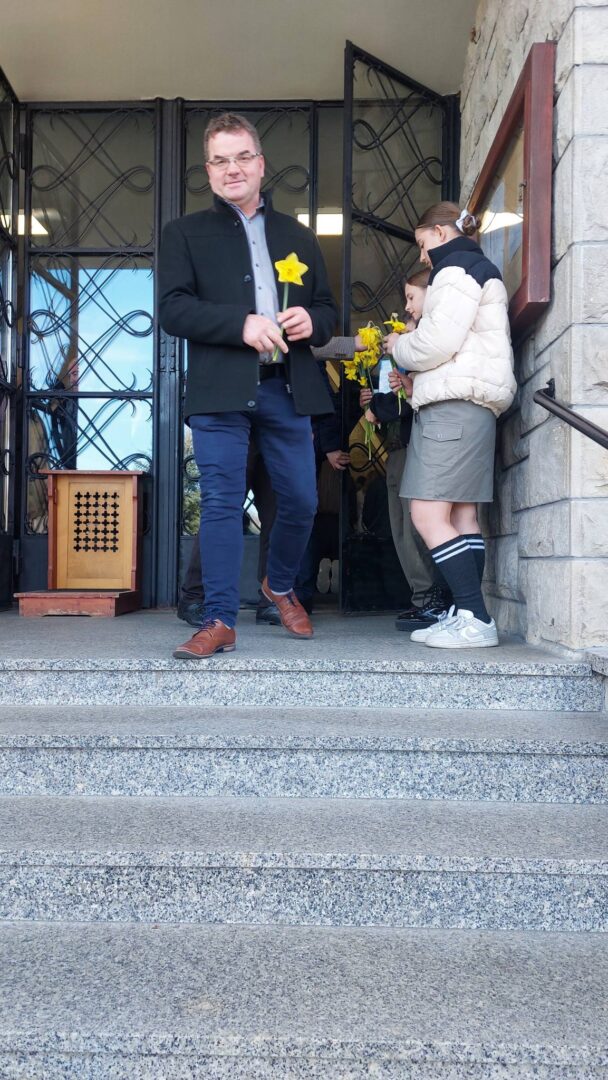 Man in a dark coat hands a bright yellow flower to a student at a doorway during a ceremony, with others nearby on the steps.