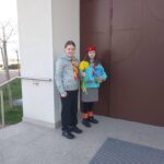 Two young girls in scouting uniforms pose outside a building, one holding a bouquet of yellow daffodils and a book.