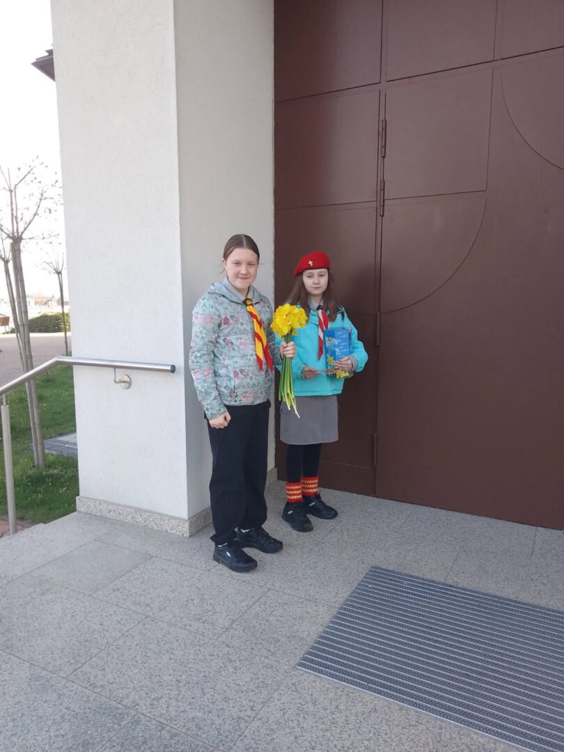 Two young girls in scouting uniforms pose outside a building, one holding a bouquet of yellow daffodils and a book.