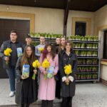 Eight people pose outside a building, each holding yellow daffodils and gift cards, in front of stacked potted plants.