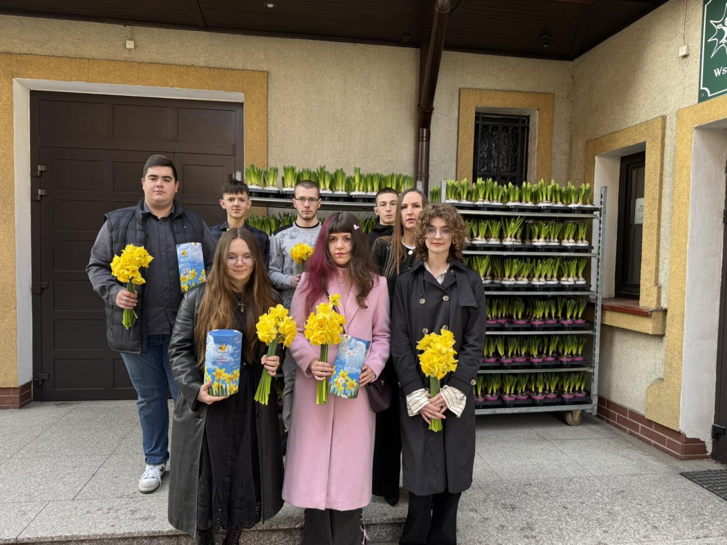Eight people pose outside a building, each holding yellow daffodils and gift cards, in front of stacked potted plants.