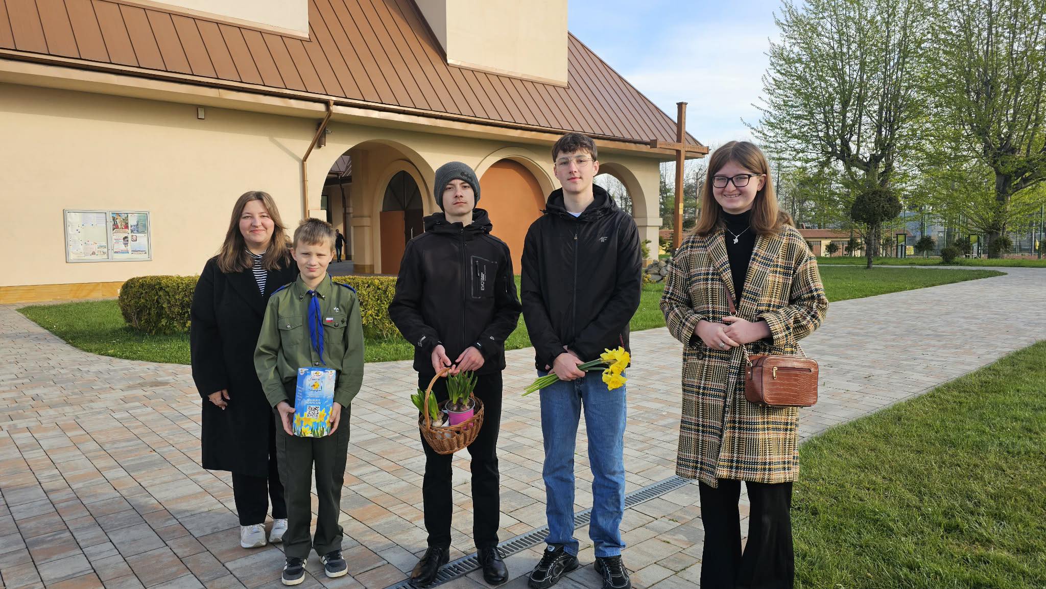 Five young people stand outside a beige building with arched entryways; two hold flowers and a wicker basket of plants.