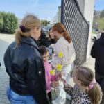 Children hand yellow daffodils to adults outside near a gate on a sunny day, with several onlookers nearby.