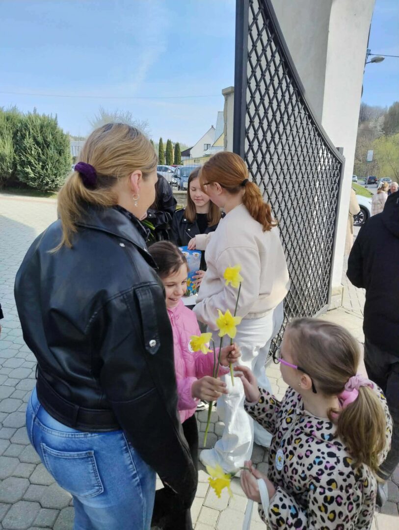 Children hand yellow daffodils to adults outside near a gate on a sunny day, with several onlookers nearby.
