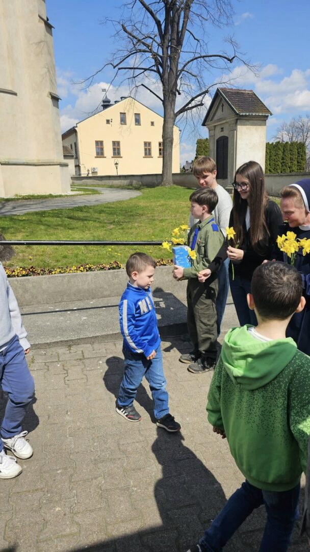 Young boy in a blue hoodie receives yellow daffodils from a group of smiling teens outdoors near a beige building on a sunny day.