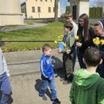 A young boy in a blue hoodie receives yellow flowers from a group of teens and kids outdoors near a park and buildings in the background.