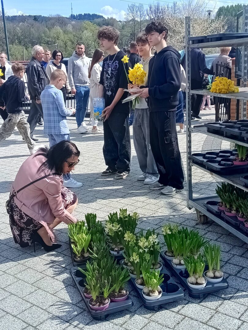 Woman crouching to inspect potted hyacinth bulbs laid out on trays at an outdoor plant market, with a crowd of people in the background and a metal shelf holding more plants nearby.
