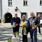 Three adults pose with flowers outdoors: yellow bouquet, potted plants, and a stone pedestal with more pots in front of a white building with barred windows.