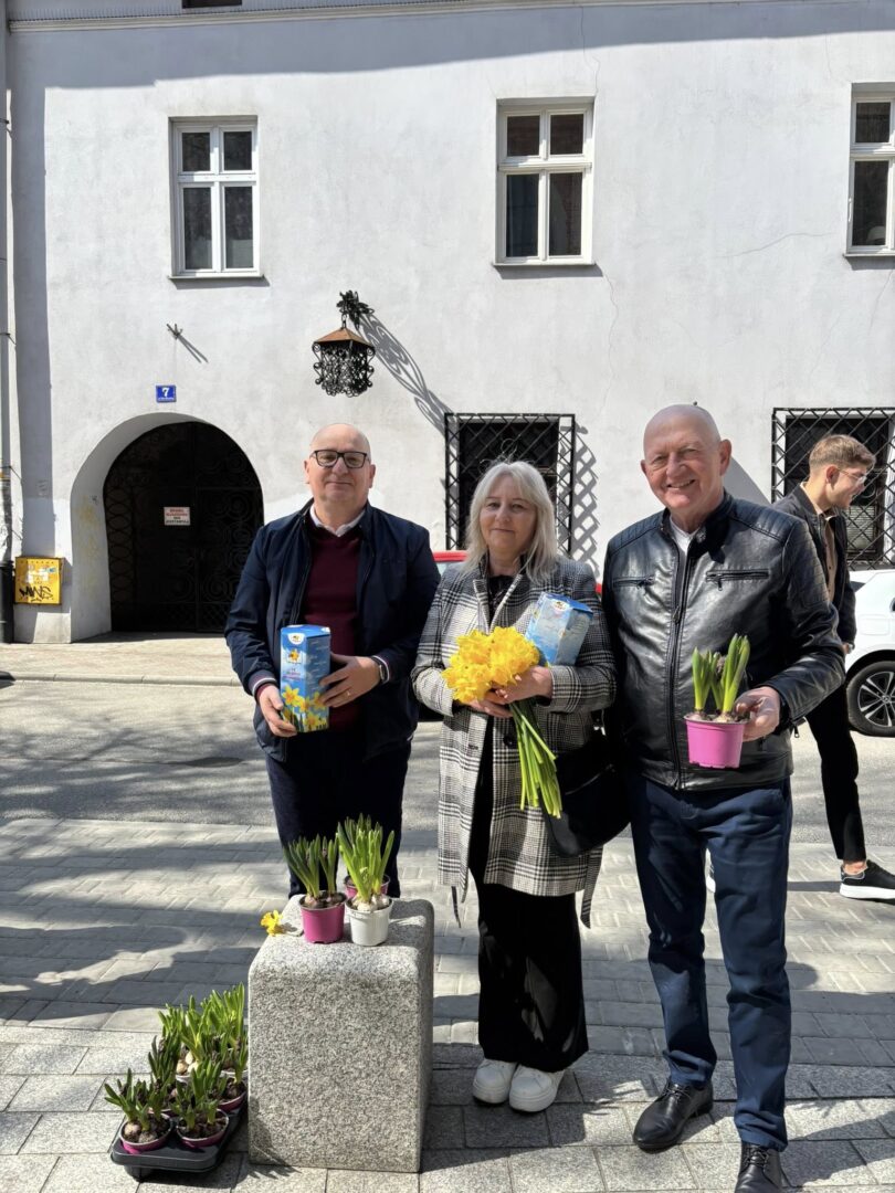Three adults pose with flowers outdoors: yellow bouquet, potted plants, and a stone pedestal with more pots in front of a white building with barred windows.