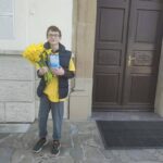 Boy standing outside a building holding a bouquet of yellow flowers and a blue card.