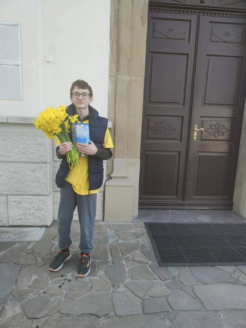 Boy standing outside a building holding a bouquet of yellow flowers and a blue card.