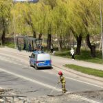 Firefighter in yellow gear sweeping debris from a road while a police van and a red fire truck are parked nearby near a bus stop by a riverbank under green trees.