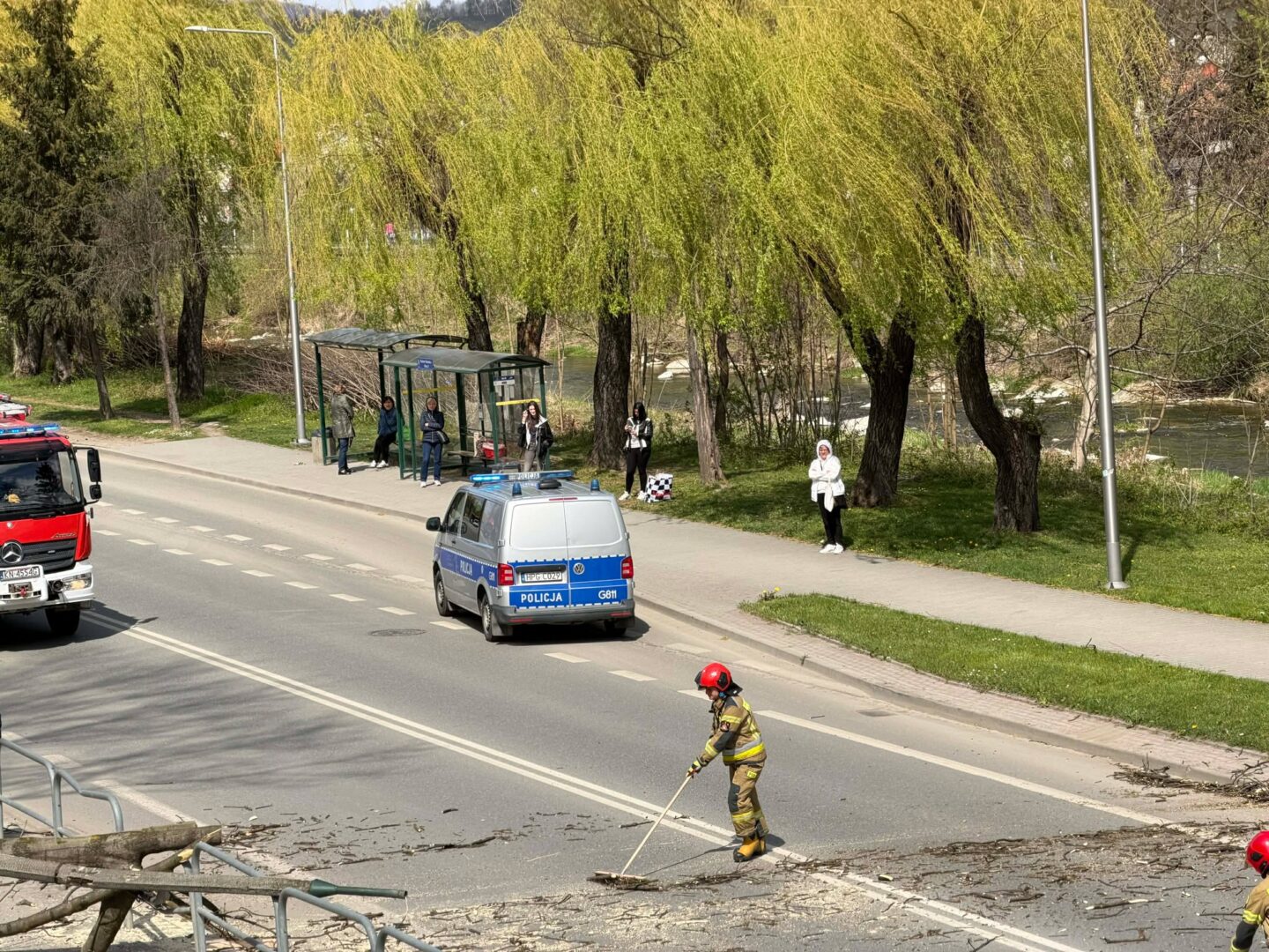 Firefighter in yellow gear sweeping debris from a road while a police van and a red fire truck are parked nearby near a bus stop by a riverbank under green trees.