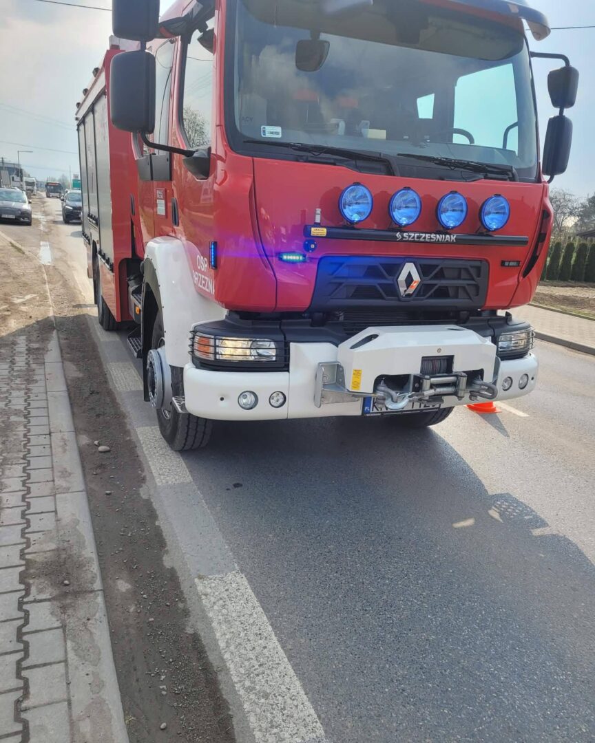 Front view of a red fire truck parked on the road, with blue emergency lights and a heavy bumper assembly visible in the foreground.
