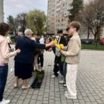 Older woman in a dark dress hands yellow daffodils to a young man in a beige jacket during an outdoor ceremony near apartment blocks; others wait in line.