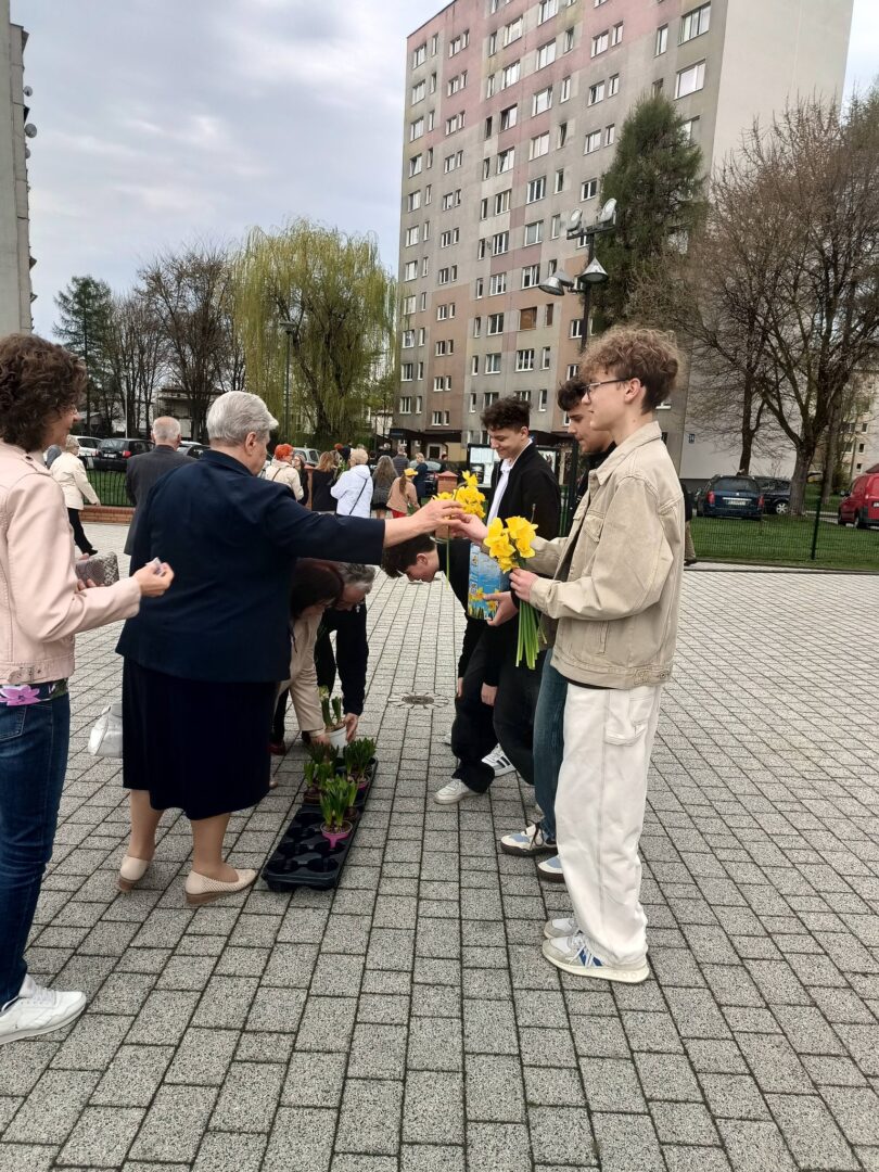 Older woman in a dark dress hands yellow daffodils to a young man in a beige jacket during an outdoor ceremony near apartment blocks; others wait in line.