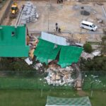 Partially collapsed green-roofed building with rubble, firefighters, and a white van at a construction site fenced area.