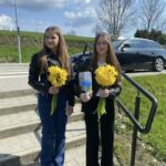 Two young women stand on a sunny sidewalk, each holding a bouquet of bright yellow daffodils, with a car and grassy area behind them.