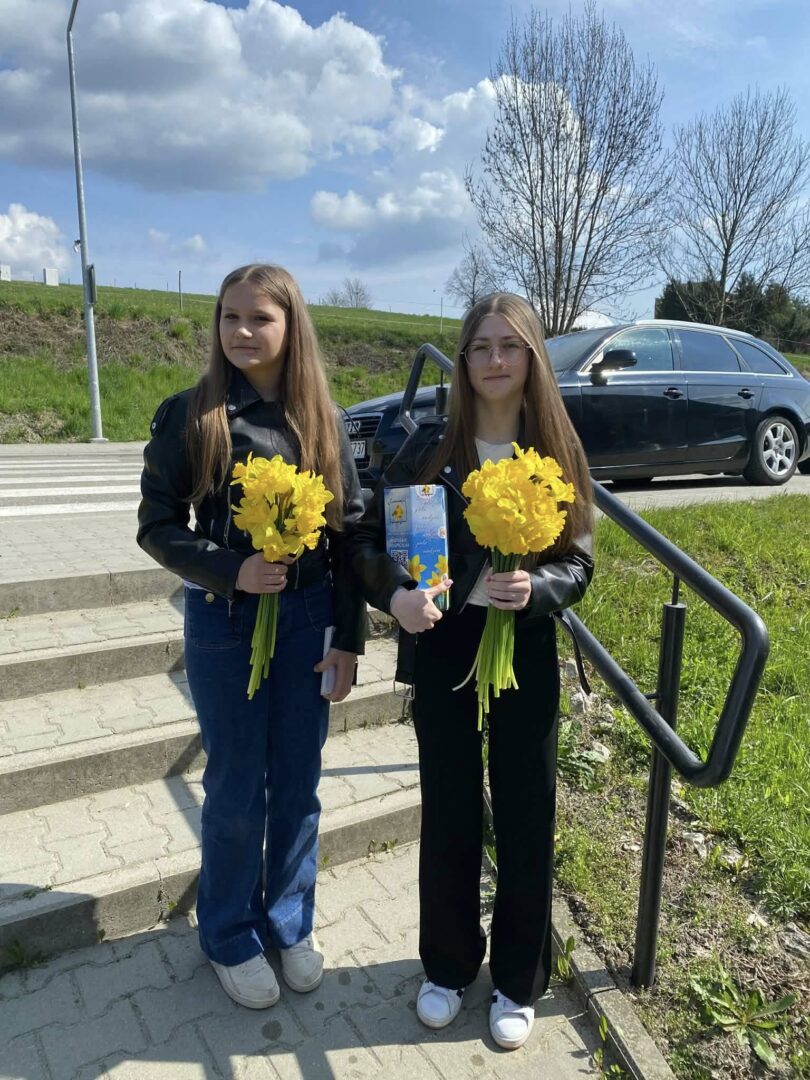 Two young women stand on a sunny sidewalk, each holding a bouquet of bright yellow daffodils, with a car and grassy area behind them.