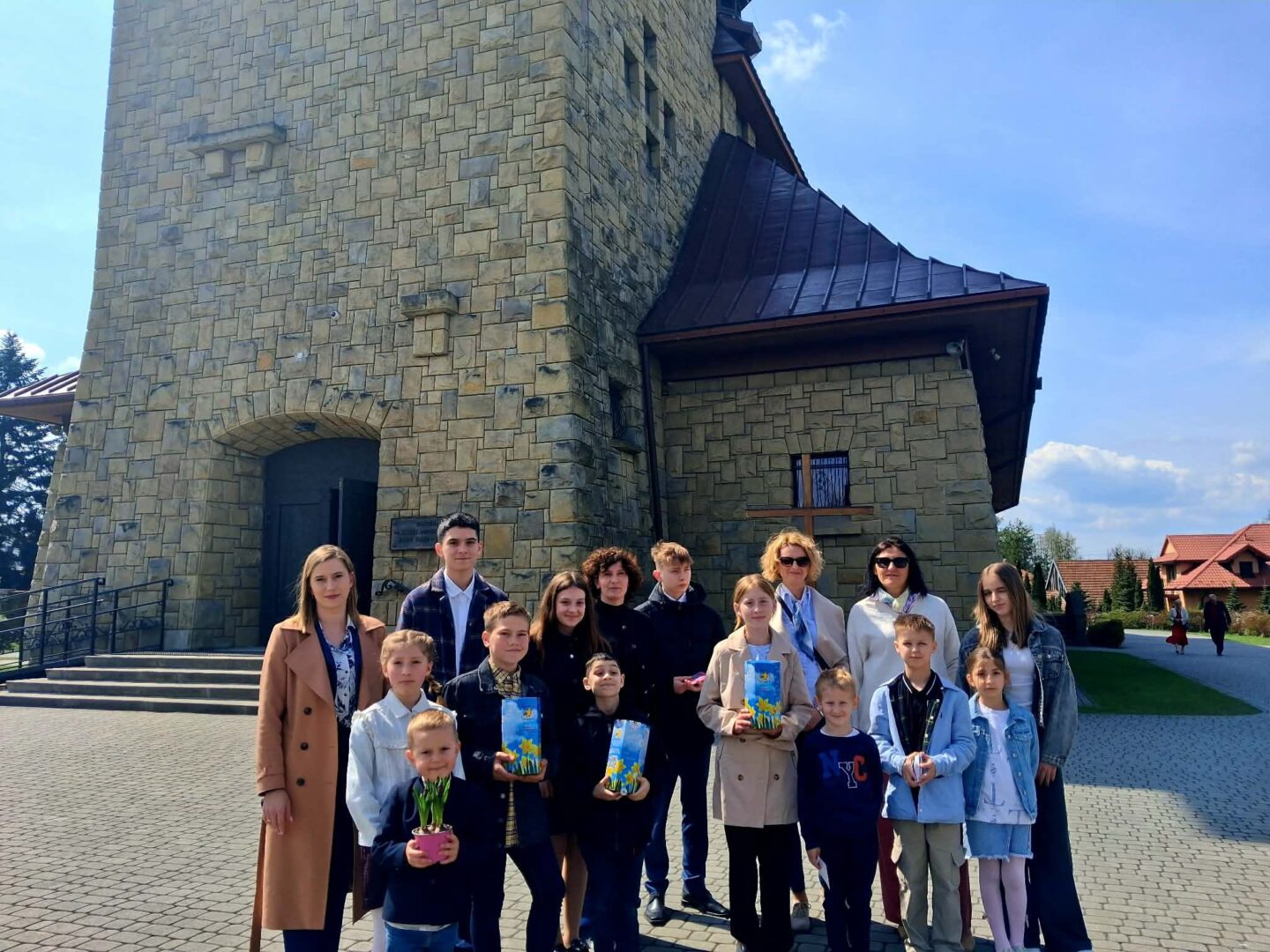 Group of children and adults posing in front of a tall stone tower with an arched doorway on a sunny day with blue sky.