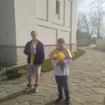 Two boys stand on a stone path beside a white building. One wears a yellow shirt and jacket and holds a book; the other, in a white robe with a yellow cape, holds a bouquet of yellow flowers (daffodils).