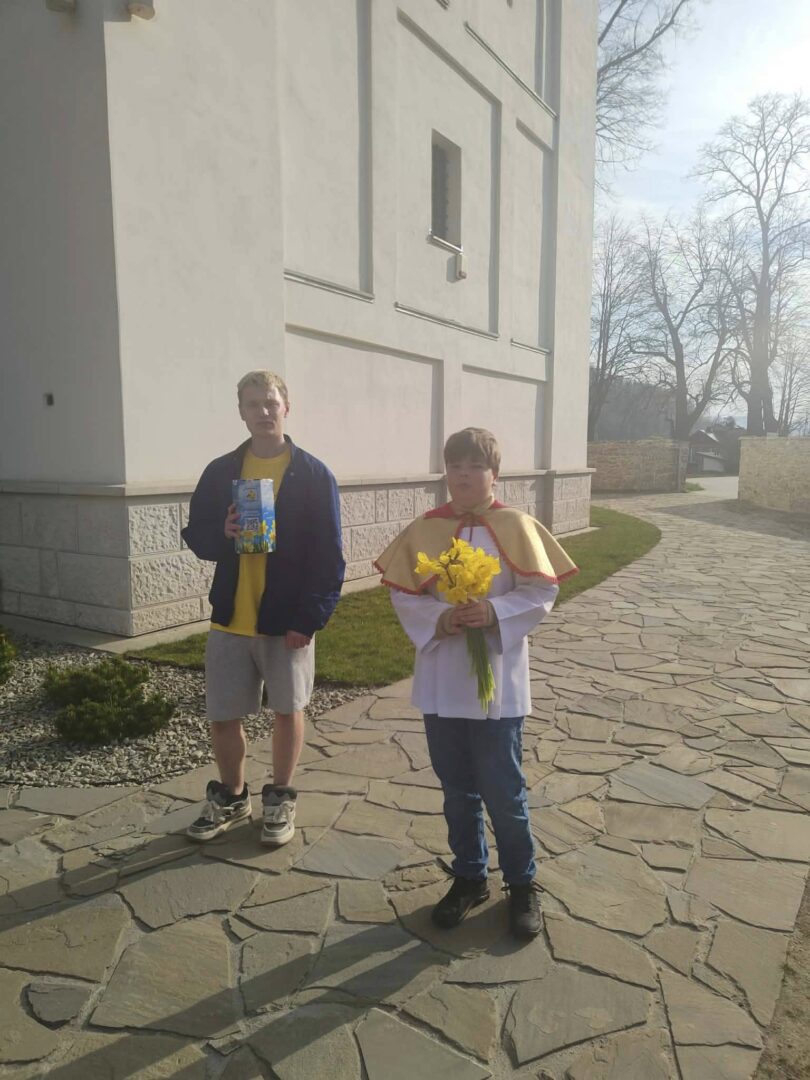 Two boys stand on a stone path beside a white building. One wears a yellow shirt and jacket and holds a book; the other, in a white robe with a yellow cape, holds a bouquet of yellow flowers (daffodils).