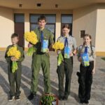 Four young scouts in uniform pose outside a beige building, each holding a yellow bouquet and a blue card; a basket of flowers sits on the ground at their feet.