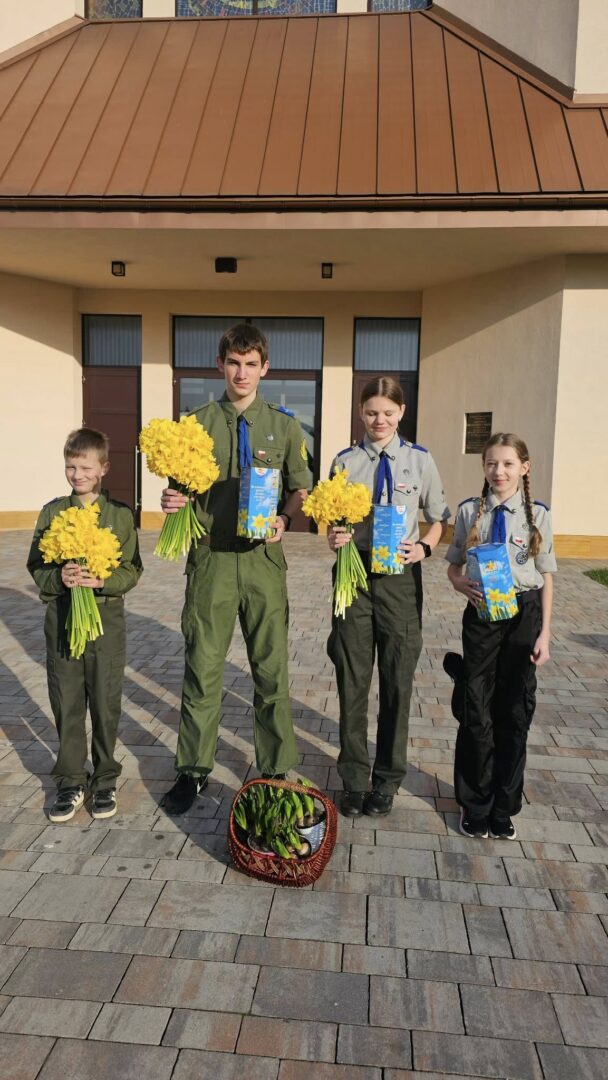 Four young scouts in uniform pose outside a beige building, each holding a yellow bouquet and a blue card; a basket of flowers sits on the ground at their feet.