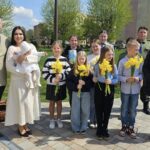 Group of adults and children outdoors in a park, many holding yellow daffodil bouquets; a woman in white cradles a baby in white.