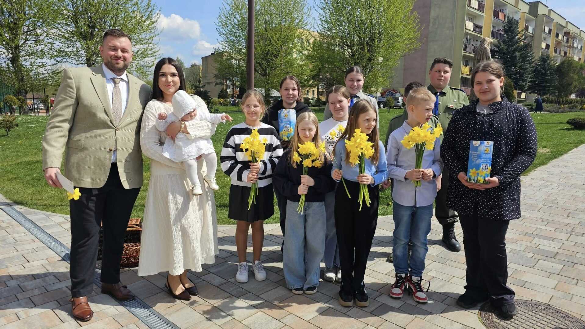 Group of adults and children outdoors in a park, many holding yellow daffodil bouquets; a woman in white cradles a baby in white.