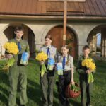 Four young scouts in uniforms stand outdoors holding yellow flowers and blue booklets, with a wooden cross behind them.