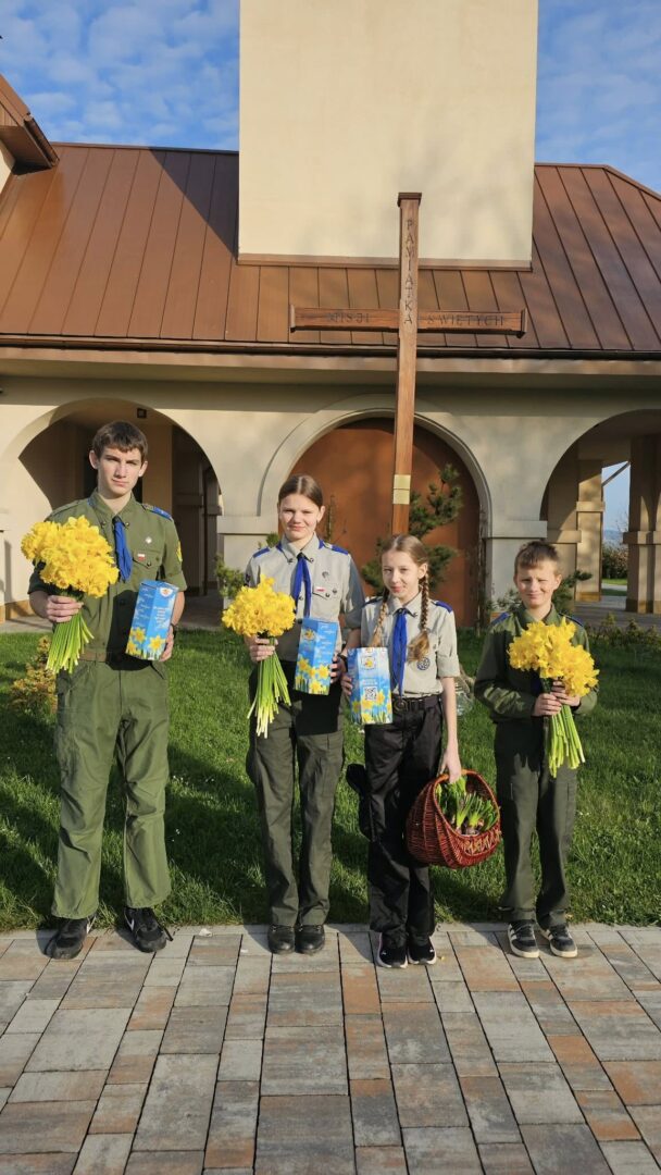 Four scouts in uniform stand outside a building with a wooden cross, holding yellow flowers and blue cards.
