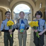 Three young scouts stand under a courtyard arch, each holding a bouquet of yellow daffodils and a blue box, smiling at the camera.