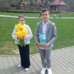 Two children stand on a paved path in a park; girl holds a bouquet of yellow flowers, boy holds a blue juice carton, both dressed nicely with a scenic village backdrop behind them.