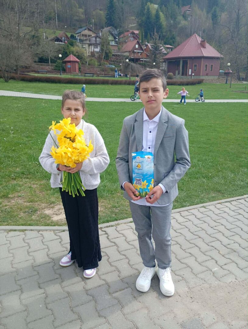 Two children stand on a paved path in a park; girl holds a bouquet of yellow flowers, boy holds a blue juice carton, both dressed nicely with a scenic village backdrop behind them.