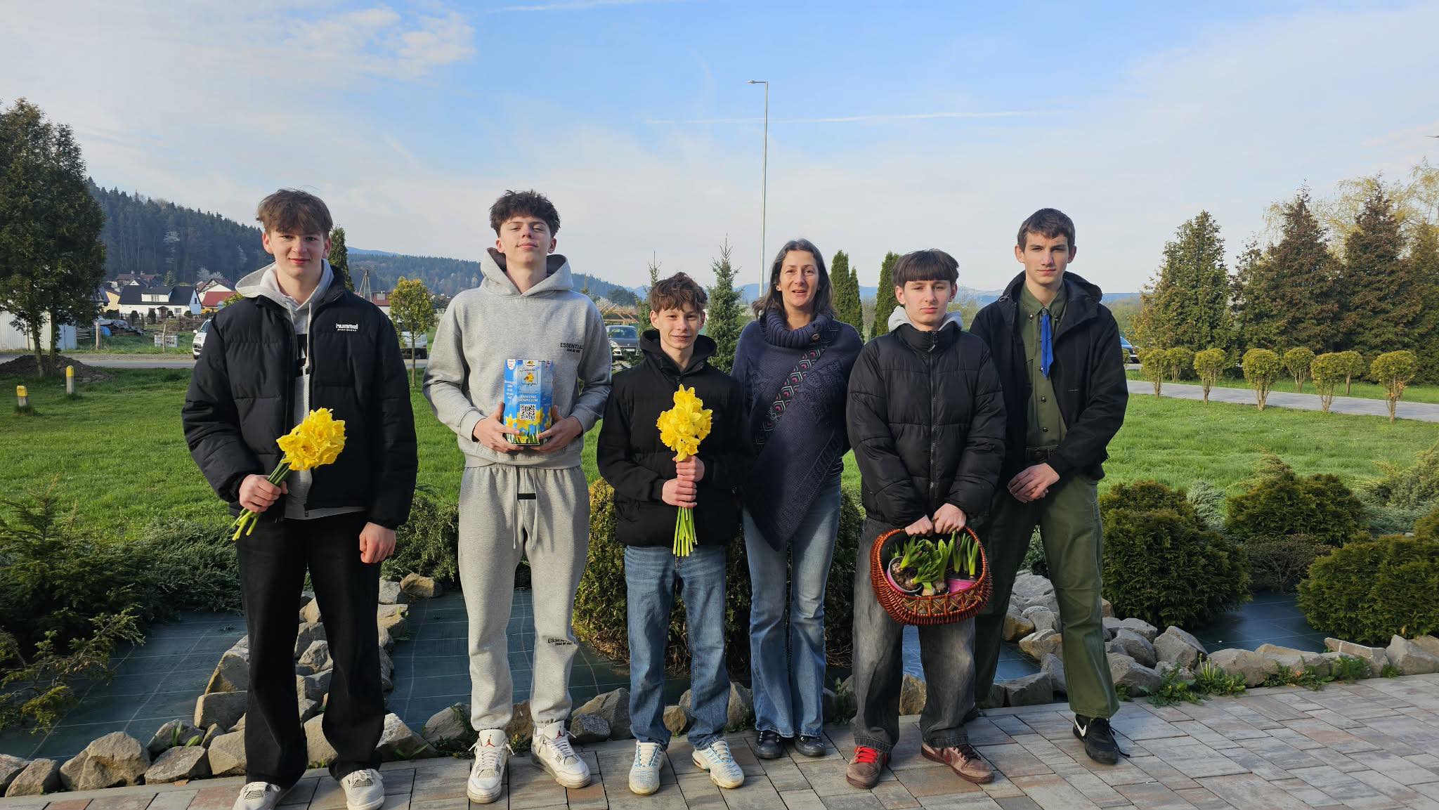 Six people stand outdoors on a paved path in a garden setting; three hold yellow flowers, one holds a gift box, and another carries a basket of plants, with an adult woman in the center-left.