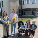 Group of five posing outdoors in front of a beige building: man holding yellow daffodils, woman with a stroller, and two kids with gift bags; smiling.