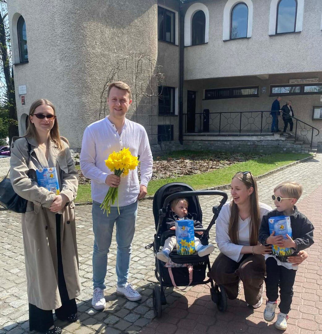 Group of five posing outdoors in front of a beige building: man holding yellow daffodils, woman with a stroller, and two kids with gift bags; smiling.
