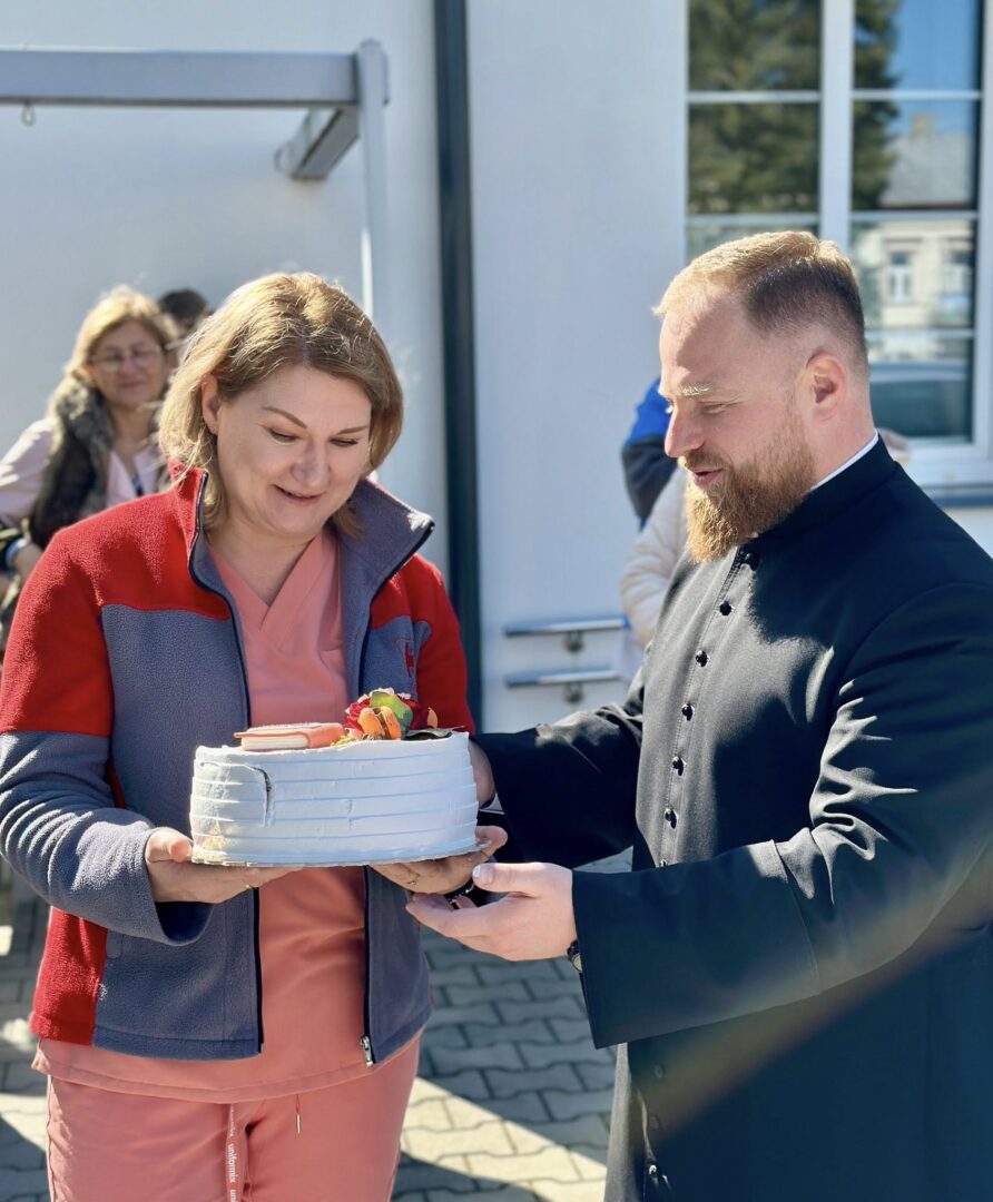 Two adults share a cake outdoors as one person hands a frosted cake to the other, fruit garnish on top.