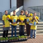 Group of six people in yellow shirts holding daffodils and gift boxes, standing on steps outside a building with potted plants nearby.