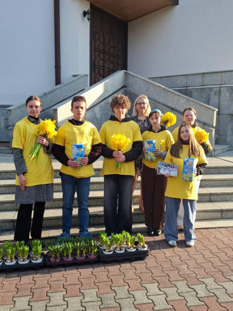 Group of six people in yellow shirts holding daffodils and gift boxes, standing on steps outside a building with potted plants nearby.