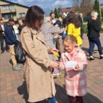 Woman in a tan trench coat hands a yellow flower to a young girl in a pink plaid coat at an outdoor gathering.