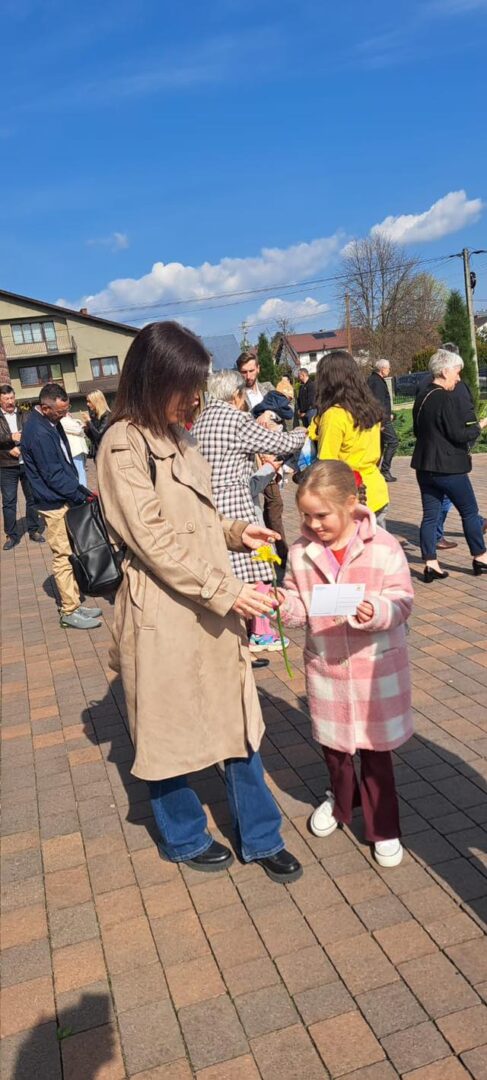 Woman in a tan trench coat hands a yellow flower to a young girl in a pink plaid coat at an outdoor gathering.