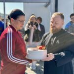 Bearded man in black clerical attire hands a white frosted cake to a woman in a red track jacket; onlookers photograph in the background outdoors.